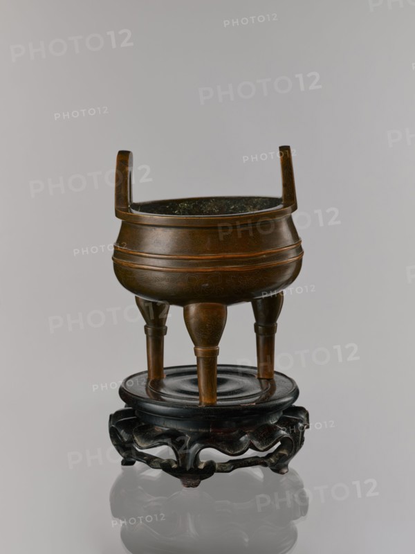 Ding incense burner on stand, Chinese, 19th century Qing, 1644–1911. Bronze censer/incense burner with decoration of lotuses, stylized hieroglyphs FU happiness, bats iconography, fo, tiger symbol, meander ornament, and one hundred antiquities baigu motif; ceremonial/ritual vessel. Round wooden turned stand on five legs; sides of the stand shaped as a double lotus rosette lower part without the top; separate upper base on foot. Dimensions: incense burner height 21 cm, diameter 15 cm; base height 7.9 cm, width 18.4 cm., A bronze tripod vessel with a rounded body and two upright handles sits on a dark carved wooden stand; the vessel shows a brownish bronze patina with subtle darker and lighter brown variations and faint engraved decorative patterns, the interior appearing darker with greenish spots, and the wooden stand is dark brown to nearly black with a glossy finish and curved ornamental cutouts.
