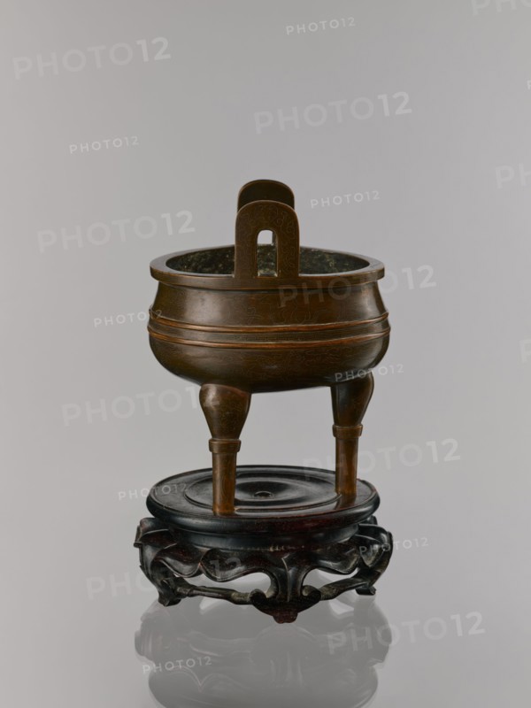 Ding incense burner on stand;, Chinese, Qing dynasty 1644–1911, 19th century. Bronze vessel censer incense burner. Decoration: lotus motifs, stylized hieroglyphs FU happiness, bats iconography, tiger symbol, fo iconographic, one hundred antiquities baigu ornament, meander ornament. Function: ritual/ceremonial vessel. Round wooden turned stand on five legs; sides shaped as double lotus rosette lower part without the top; separate upper base on foot. Dimensions: incense burner height 21 cm, diameter 15 cm; base height 7.9 cm, width 18.4 cm., A brown metal censers with a rounded body and a pair of vertical handles sits on a dark brown wooden stand with carved openwork legs; the censer has a polished surface with subtle engraved patterns and rests on three tapered legs above a circular top on the stand, and the visible colors include brown and dark brown against a light gray background.