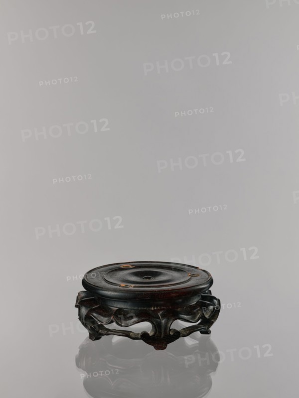 Ding incense burner on stand, Chinese, Qing dynasty 1644–1911. Incense censer ritual vessel, 19th century. Round wooden turned stand on five legs; sides shaped like double lotus rosette lower part without the top; upper base on foot separate. Decoration: lotus motifs, stylized hieroglyph FU happiness; iconography includes bats, fo, tiger, lotuses, one hundred antiquities baigu, meander ornament. Dimensions: incense burner height 21 cm, diameter 15 cm; base height 7.9 cm, width 18.4 cm., A low, round wooden stand with a dark brown to almost black finish sits on a reflective surface, showing a subtle reflection beneath it; the top surface is slightly concave with a central shallow depression and a few small areas where the finish is worn to a lighter brown, while the carved base features flowing, curving cutouts and scrolled feet in the same dark brown hue with some lighter brown highlights.