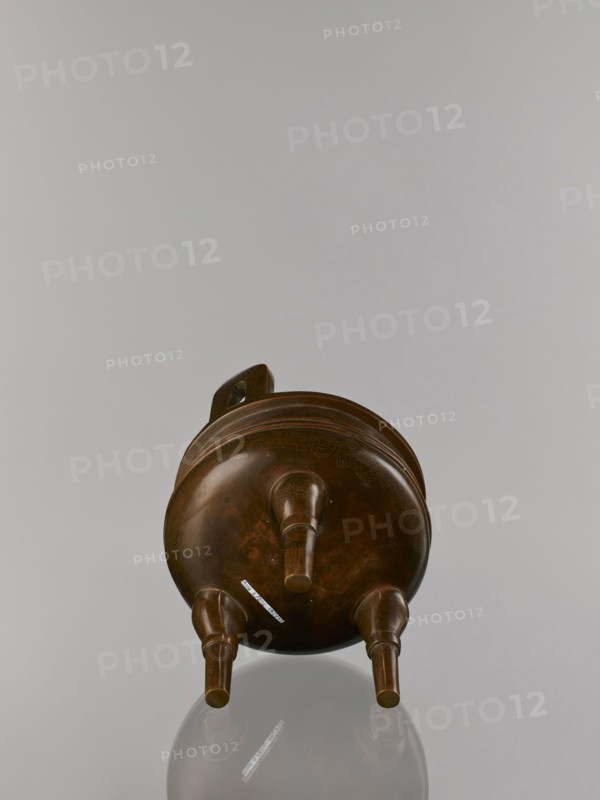 Ding incense burner on stand, maker, Chinese, Qing dynasty 1644–1911; 19th century. Bronze vessel censer/incense burner with decorations of lotuses, bats iconography, fo iconography, tiger symbol, meander and one hundred antiquities baigu ornament; stylized hieroglyph FU happiness. Round turned wooden stand on five legs; sides shaped as double lotus rosette lower part without the top; separate upper base on foot. Dimensions: burner height 21 cm, diameter 15 cm; base height 7.9 cm, width 18.4 cm., A brown rounded vessel with a handle on its upper side rests on three short cylindrical legs, showing a smooth polished surface with subtle darker brown variations and faint decorative incised lines; the background is a pale gray and the vessel casts a soft reflection below it.