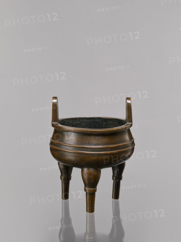 Ding-type incense burner on a stand, Qing dynasty 1644–1911, 19th century, China. Bronze ceremonial vessel decorated with lotus motifs, stylized FU happiness hieroglyphs, lotuses, bats, fo, tiger, meander ornament, and the one hundred antiquities baigu motif. Round wooden turned stand with five legs on a pedestal shaped like a lotus rosette. Function: ritual/ceremonial incense burner. Dimensions: incense burner height 21 cm, diameter 15 cm; base height 7.5 cm, width 18.4 cm., A bronze tripod vessel with a round body and a dark interior sits against a pale gray background; the vessel has a warm brown and dark bronze patina with subtle etched patterns on its surface, a flared rim supported by two short upright handles, and a ringed band encircling the upper and midsection, standing on three tapered legs that reflect faintly on the glossy surface below.