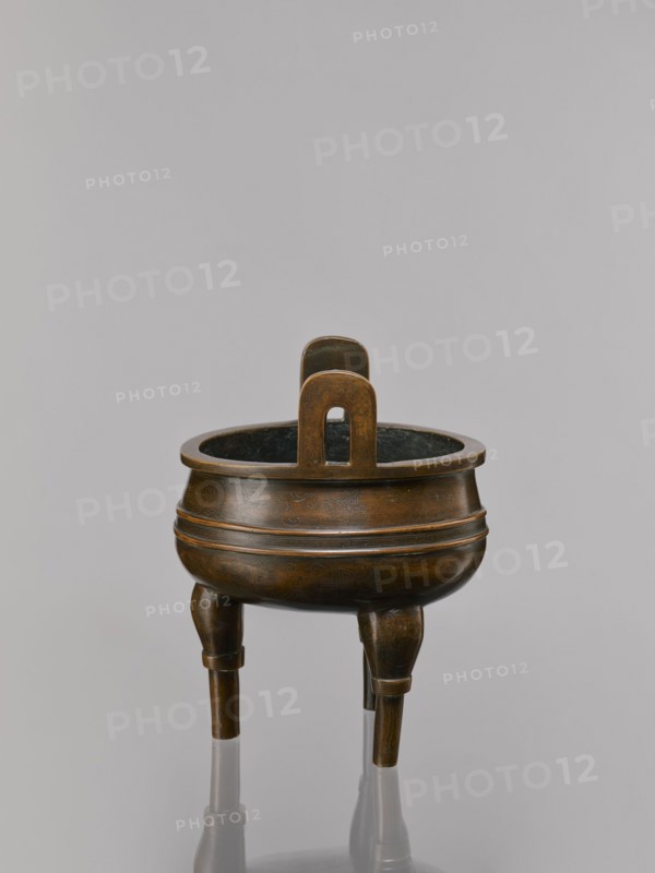 Ding-type incense burner on a stand;, 19th century, China, Qing dynasty 1644–1911. Bronze ceremonial ritual vessel incense burner decorated with lotus motifs, bats iconography of happiness, fo, tiger symbol, meander and one hundred antiquities/baigu ornament, stylized hieroglyphs reading FU happiness. Round turned wooden stand with five legs on a pedestal shaped as a lotus rosette. Incense burner: height 21 cm, diameter 15 cm. Stand base: height 7.5 cm, width 18.4 cm., A bronze tripod vessel with a rounded body and a concave rim sits against a neutral gray background, showing a warm brown and dark bronze patina with subtle greenish-black interior tones; the vessel rests on three slightly tapered legs that reflect on the surface below, and a vertical handle with a rounded top and an arched cutout rises from the rim, all surfaces displaying faint engraved patterns and a smooth, aged finish.