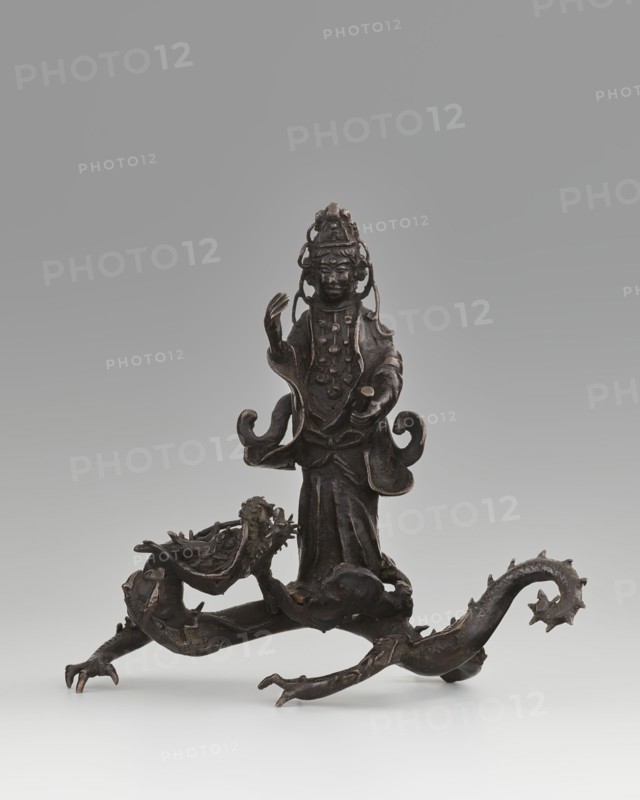 Guanyin on a dragon;, Chinese; Qing dynasty 1644–1911, 18th century; sculpture, figurine; materials not specified; subject: Guanyin bodhisattva of mercy riding a dragon with a pearl; iconography: dragons, Buddhism, goddesses; dimensions: overall height 15 cm, width 7.1 cm; dragon element height 4 cm, width 8.3 cm., A dark bronze figurine stands on a sculpted base formed by a sinuous dragon-like creature, with a few curling limbs and clawed feet extending outward. The standing figure wears layered garments with flowing sleeves and rounded ornamental details on the chest, and a tall, elaborate headdress with looping elements that frame the face. One arm is raised with an open hand while the other extends forward holding a small cylindrical object. The dragon-like base has a textured head with an open mouth and ridged spine, and a curled tail with pointed projections. The visible colors are dark brown and black tones with subtle lighter highlights where the metal catches light, set agains...