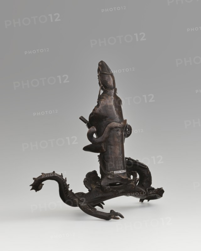 Guanyin on a dragon; creator, ; Chinese; Qing dynasty 1644–1911; Buddhism; bodhisattva/goddess of mercy; dragons iconography, dragon with a pearl; 18th century, China; sculpture, figurine; overall height 15 cm, width 7.1 cm; dragon: height 4 cm, width 8.3 cm., A dark brown sculptural object with a glossy surface stands on a pale background, featuring an upright central form that tapers toward the top and a looping element wrapped around it. The base extends into a sinuous, dragonlike appendage with ridged and spiky textures, clawed feet, and curved tail, all sharing the same dark brown tone. Subtle highlights of lighter brown and small areas of muted sheen appear where the surface catches light, and the surrounding space is a smooth gradient of pale gray to off-white.