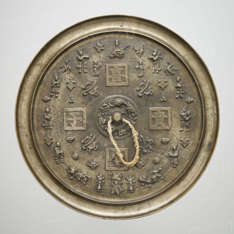 Mirror, gift, Chinese, Qing dynasty 1644–1911. Bronze mirror with polished obverse; reverse contains characters with good wishes and Chinese symbols for happiness associated with Taoism. 19th century. Mirror, diameter 34.5 cm, height 1.5 cm, weight 4.015 kg., A round metal mirror with a raised rim and a central boss, shown from above, with an attached braided loop of light beige cord; the mirror surface is an aged bronze color with darker brown areas and greenish patina; the surface is decorated with a ring of small raised figurative and abstract motifs including a few humanlike figures, a few animallike shapes, and a few knotlike designs arranged around the center, plus a few square framed panels containing raised shapes; the rim and outer edge have a slightly brighter golden-bronze tone while the decorative reliefs show varied brown and shadowed tones.