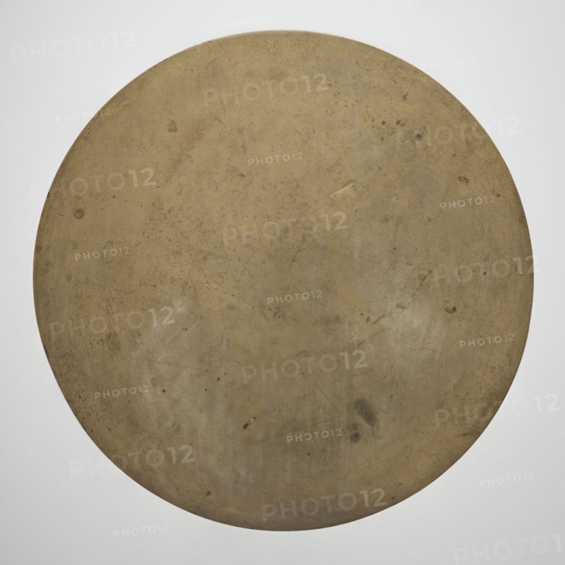Mirror, Paderewski, Ignacy Jan 1860-1941, Chinese art, Chinese culture, Qing 1644–1911, Chinese symbols, happiness symbol, Taoism, characters on the reverse with good wishes. Obverse polished. 19th century, China. Mirror; weight 4.015 kg; height 1.5 cm; diameter 34.5 cm., A large circular flat object fills the frame, showing a worn, mottled surface in shades of brown, tan, and muted gold with areas of darker brown and faint greenish spots; the surface has subtle scratches, scuffs, small stains, and irregular patches of lighter and darker tones, while the background around the circle is a smooth pale gray.