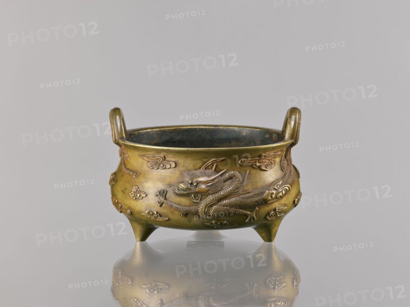Li-type incense burner censer, Chinese, 18th century copy after a 15th-century original, Qing dynasty 1644–1911. Bronze vessel, height 11.5 cm, width 17.6 cm. Decoration: two dragons among clouds; motifs include dragons and clouds. Ceremonial/ritual ding-style vessel; backdated signatures., A small bronze tripod vessel with a round bowl and two upright loop handles, resting on three short conical legs, shown against a plain light gray background; the vessel surface is a warm golden-bronze with darker brown and greenish patina in places, and it is decorated with raised dragon motifs and stylized cloud-like forms in the same metal finish; the interior appears darker with a bluish-green tinge, and the vessel casts a soft reflection on the glossy surface beneath it.