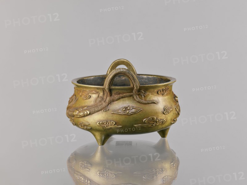 Li-type incense burner, ding vessel form, Chinese art; dragons and clouds iconography; ceremonial/ritual vessel; Qing dynasty 1644–1911, 18th century copy after a 15th-century original; backdated signatures; Decoration: two dragons among clouds; vessel censer; height 11.5 cm, width 17.6 cm., A small rounded metal vessel with a warm golden-bronze color sits on a reflective surface, showing a faint mirrored image beneath; the vessel has a wide rim and a slightly darkened interior, and rests on three short pointed legs. A textured handle arches over the top, and a raised snake or dragon-like figure wraps around the body, rendered in the same golden-bronze tone with fine scale details. The exterior surface is decorated with several low-relief cloud-shaped motifs in matching bronze, and the overall background is a smooth, pale gray.