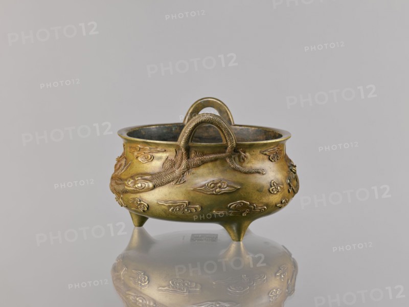 Li-type incense burner censer, Ding-style vessel, Chinese, 18th century copy after a 15th-century original, Qing dynasty 1644–1911. Bronze censer, height 11.5 cm, width 17.6 cm. Decoration: two dragons among clouds; backdated signatures; ceremonial/ritual function., A small metal vessel with a rounded body and three short conical legs sits on a reflective surface, showing a mirrored image beneath. The vessel is primarily brass-colored with areas of darker patina and greenish tones inside the rim. Raised decorative elements in the same metallic hues cover the exterior, including a textured rope-like handle arching over the top and a sculpted long animal motif wrapping around the body. The background is a smooth light gray, and the reflection on the surface echoes the vessel's brass and darker greenish colors.