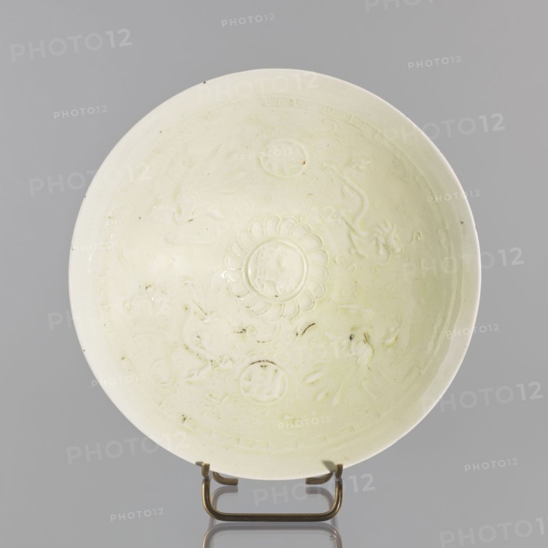 Rice bowl, Yuan dynasty 1271–1368, late 13th century, China; ceramic vessel decorated with rosettes, dragons and phoenixes fenghuang; height 6.5 cm, diameter 19.8 cm; used for rice., A shallow round dish with a pale creamy off-white glaze displayed upright on a small metal stand, showing a central raised floral medallion surrounded by shallow embossed decorative motifs and curved ornamental designs, with subtle variations in glazing tone and a few small darker specks and spots on the surface, set against a neutral gray background; the metal stand is a brass-like color.