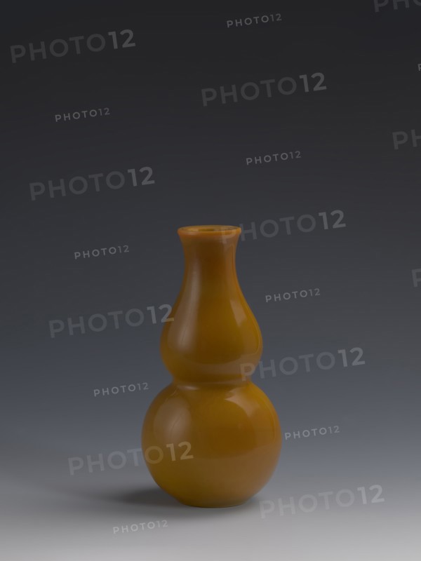 Vase, Chinese, Qing dynasty 1644–1911, 18th century. Double gourd-shaped vessel made of yellow glass, cast and cut, opaque; stained glass effect. Height 16.5 cm; diameter 8.0 cm; weight 0.41 kg., A smooth, glossy gourd-shaped vase in a warm amber color sits centered on a surface, showing reflective highlights and subtle shadows; the background transitions from a dark gray at the top to a lighter gray near the bottom, and the surface beneath the vase is a pale gray with a soft shadow on one side.