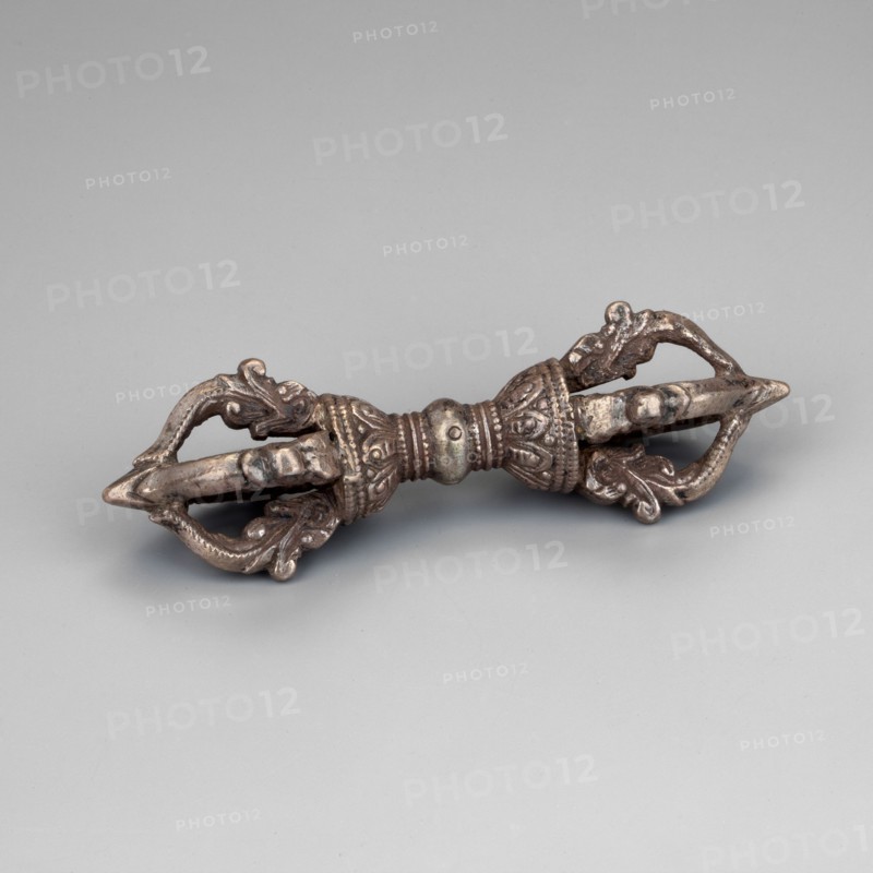 Vajra;, Himalayan, Tibetan Buddhist art; vajra ceremonial object; 18th century ; Tibet ; height 9.5 cm., The image shows a metallic object with an elongated symmetrical form resting on a flat surface; it has ornate sculpted sections with curved, leaflike and scrolllike decorative elements at both ends and a central banded, beaded area. The object appears to be made of a dark silvery metal with areas of darker patina and hints of brownish tones. The background and surface are a smooth, pale gray.