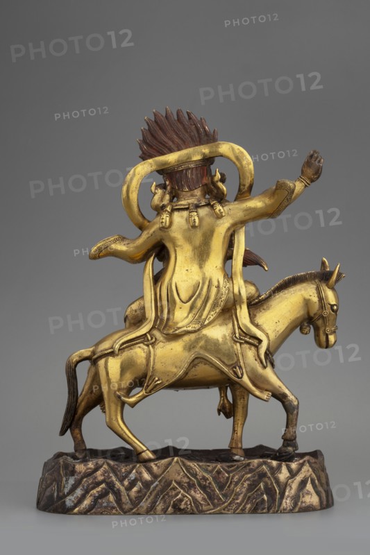 Palden Lhamo, unknown sculptor, Tibetan Buddhist art, Himalayan art, Himalayan culture, dharmapala iconography, mules, skins human, tiaras, skulls, 18th century, Tibet, sculpture, figurine, height 16.7 cm, weight 1.11 kg, A gilded metal figure of a person seated on a horse, shown from the back, with a flowing robe and an ornate sash draped over shoulders and down the rider’s back; the rider has a tall, spiked hairpiece in a darker brown tone and a rounded halo-like element behind the head. The horse and rider are both gold in color with darker brown accents on the hairpiece, tail, hooves, and base. The horse stands on an irregular textured base with a mottled gold and dark brown surface that suggests a rocky ground. The rider’s raised arm and the sculpted folds of the robe are clearly visible, as are small decorative details on the rider’s sleeves and the horse’s bridle. The background is a smooth, neutral gray.