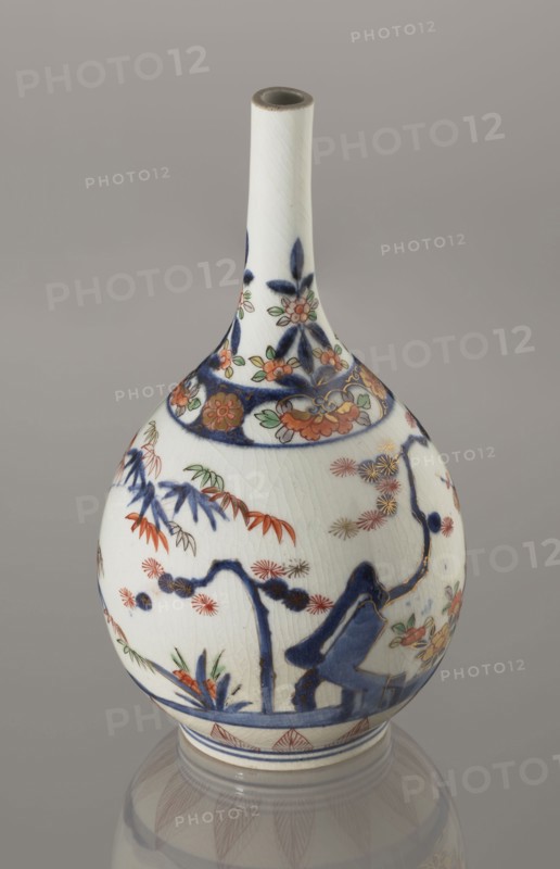 Bottle, ca. 1700–1730, porcelain; Arita, Japan. Height 22.3 cm, diameter 12.2 cm. Decorated in Imari palette with motifs of bamboos, pines, and plums — the Three Friends of Winter — associated with sake vessels; Japanese ceramicist/author unknown., A ceramic vase with a long narrow neck and a bulbous body, shown against a plain gray background. The vase is primarily white with visible crackle glazing and is decorated with painted floral and botanical motifs in dark blue, orange, green, red, and gold. The neck features dark blue leaf-like shapes and small orange and green flowers, while the rounded body displays dark blue branches and stylized foliage, orange and red flowers, green leaves, and gold-accented blossoms. A dark blue band encircles the lower body above a narrow foot ring, and the base reflects softly on the surface below, showing muted gray and the vase’s painted patterns.