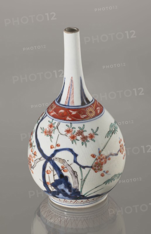 Bottle, early 18th century, Japan Edo, 1603–1868. Porcelain sake bottle decorated in the kakiemon style with a motif of the three friends of winter bamboo, plum, and pine; rock. Also features two borders with plant motifs and floral patterns on the neck. Height 22.1 cm; diameter 12 cm., A white ceramic vase with a long narrow neck and rounded body sits against a neutral gray background, decorated with floral and branch motifs in red, green, and blue, including red blossoms with green leaves and thin black branch lines, a band of red with floral scrollwork near the neck, and bold blue brushstrokes forming abstract shapes; the vase shows fine crackle texture in the glaze and a faint reflection beneath it on the surface.