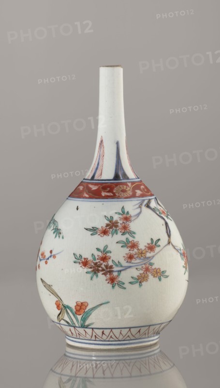 Bottle, Edo period 1603–1868, early 18th century, Japan. Porcelain sake bottle vessel, height 22.1 cm, diameter 12 cm. Decorated in the kakiemon style with a motif of the three friends of winter bamboo, plum, and pine; rock, and two borders with plant motifs and floral patterns on the neck. Materials: ceramics porcelain., A pale ceramic bottle with a long narrow neck and bulbous body sits against a neutral gray background; the surface is primarily white with painted floral motifs in red, green, blue, and touches of yellow, showing branches with clusters of red-orange blossoms and green leaves, a band of red with floral ornamentation near the shoulder, blue linear accents around the base and neck, and a small area of brownish discoloration near the top rim.