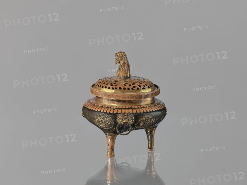 Incense burner kôro of the ding type with a cover. Incense burner with two handles knobs, on three legs. Openwork cover decorated with a floral tendril and topped with a komainu figure mythical beast. Meiji period, 2nd half of the 19th century 1868–1912. Japan. Bronze, height 13.8 cm, diameter 12.9 cm., The image shows a small ornate tripod vessel with a perforated lid topped by a sculpted animal figure; the vessel sits on a reflective surface against a plain background. The object displays metallic colors including gold, bronze, and dark brown, with a few areas of lighter worn metal; the lid has a patterned perforation and a textured rim, the body features decorative raised medallions and a ring hanging from one side, and the three tapered legs show wear and metallic sheen.