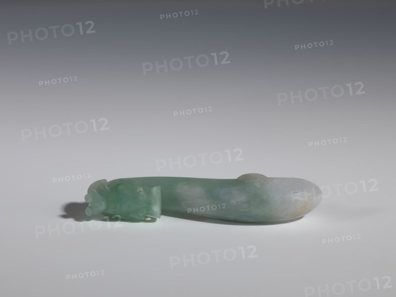 Belt clasp, purchase provenance. Chinese, Qing 1644–1911. Green-gray jadeite, semi-transparent with slight colloid clusters. Full-plastic carved head with dragons iconography. Smooth handle. Function: belt buckle for men's clothing, ornament. 17th–18th century. Dimensions: height 2.0 cm, width 9.1 cm, depth 1.7 cm., A smooth, elongated carved object lies horizontally on a light surface against a gradient gray background; the object is primarily green with areas of pale white and subtle brownish discoloration, and one end features a carved decorative form with rounded protrusions while the other end is rounded and smooth, casting a soft shadow beneath.