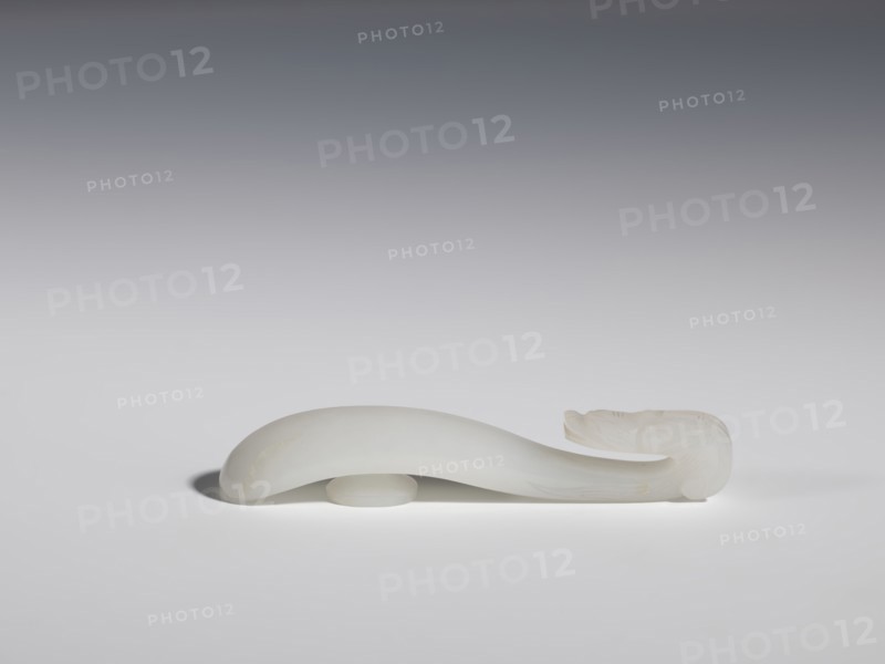 Belt clasp, Chinese, dragons iconography, Qing 1644–1911, handle devoid of engraved decoration, polished smoothly. Milky-white jadeite, slightly translucent. Full-plastic carved head, engraved, 18th–19th century, China, belt buckle, height 1.5 cm, width 8.8 cm, depth 1.7 cm, A smooth, curved pale object with a translucent white appearance rests on a flat surface, showing a rounded, thicker midsection and a tapered end that curls upward into a more detailed carved shape; the background is a gradient from dark gray at the top to light gray near the surface, and the object casts a soft shadow on the surface.