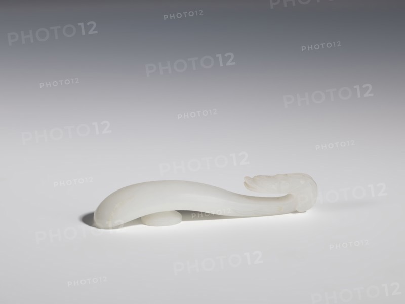 Belt clasp, Chinese, dragons iconography, Qing 1644–1911, handle devoid of engraved decoration, polished smoothly. Milky-white jadeite, slightly translucent. Full-plastic carved head, engraved, 18th–19th century, China, belt buckle; height 1.5 cm, width 8.8 cm, depth 1.7 cm., A smooth, curved object made of a translucent white material rests horizontally on a flat surface, casting a soft shadow beneath it; the object tapers at one end and has a slightly bulbous, rounded opposite end with subtle carved ridges and contours; the background transitions from medium gray at the top to pale gray near the surface, and the visible colors are white, gray, and faint cream tones on the object.