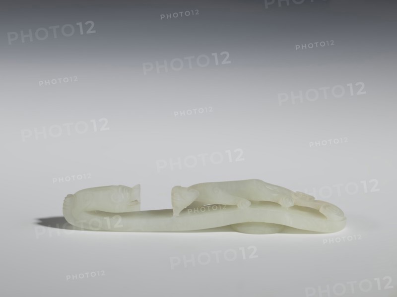 Belt clasp, Chinese culture, dragons iconography, lizards zool., Qing 1644–1911. The head is in the shape of a two-horned dragon's head with an open mouth, and the handle features a lizard. Milky-white jadeite, slightly translucent with slight colloid clusters. Carving, engraving; 18th–19th century, China. Belt buckle; height 2.1 cm, width 12.8 cm, depth 2.2 cm., A pale, creamy white carved object rests on a smooth surface against a gradient gray background, shaped like a long, sinuous form with rounded ends and subtle raised details suggesting limbs and facial features, casting a soft shadow beneath it; the overall visible colors are creamy white, light gray, and darker gray in the background.