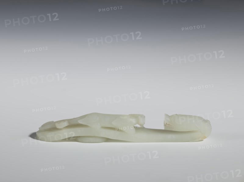 Belt clasp;, Chinese, Qing dynasty 1644–1911, 18th–19th century. Milky-white jadeite, slightly translucent with slight colloid clusters; carved and engraved. The head is in the shape of a two-horned dragon's head with an open mouth, and the handle features a lizard. Belt buckle; height 2.1 cm, width 12.8 cm, depth 2.2 cm., A pale, translucent carved object rests horizontally against a neutral background, showing a smooth, milky white surface with subtle areas of pale yellow and cream. The carving features flowing, elongated forms with rounded contours and gentle hollows, including a raised central section and curved end pieces that suggest stylized heads or decorative terminals. Soft shadows beneath the object create a sense of low elevation, and the overall palette includes shades of white, off-white, cream, and faint yellow against a gradient background ranging from light gray to darker gray at the top.
