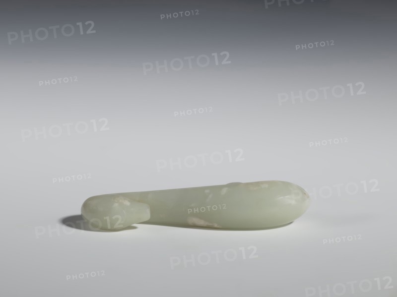 Belt clasp, Chinese, Qing 1644–1911, 17th–18th century. Milky-white slightly translucent jadeite, smooth rounded handle. Full-sculpture carved lizard head, engraved. Belt buckle. Height 2.2 cm, width 10.5 cm, depth 2.1 cm., A smooth, elongated object rests horizontally on a flat surface, appearing to be made of a pale, translucent material in shades of light green and off-white with some faint cloudy patches. The object has rounded ends, one end slightly bulbous and the other tapering to a smaller rounded tip, and it casts a soft shadow on the surface beneath it. The background transitions from a darker gray at the top to a lighter gray near the surface, and the overall lighting is soft and diffuse, highlighting the object's glossy surface and subtle color variations.