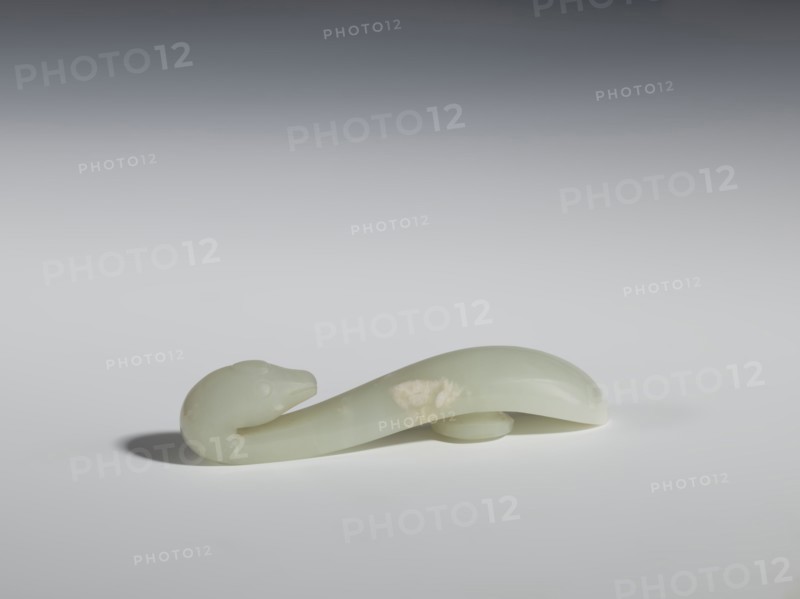 Belt clasp, Chinese, lizards zoological motif, Qing 1644–1911. Smooth, rounded handle. Milky-white jadeite, slightly translucent. Full-sculpture carved head, engraved. 17th–18th century, China. Belt buckle. Height 2.2 cm; width 10.5 cm; depth 2.1 cm., A smooth, curved object resembling an elongated animal form rests on a flat surface, showing a rounded head with subtle facial contours and a tapering body that loops gently; the object appears to be made of a polished, translucent material with a pale green color and a few slightly lighter patches, casting a soft shadow on the surface beneath, with the background transitioning from light gray near the surface to darker gray toward the top.