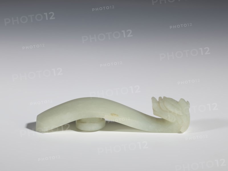 Belt clasp, stones, Milky-white jade with colloid clusters. Full-plastic carved head. Chinese, Qing dynasty 1644–1911, 18th–19th century. Dragons iconography. Belt buckle. Height 2.1 cm; width 10.1 cm; depth 2.2 cm., A pale green, smooth carved object with a curved elongated body resting on a round base and ending in a stylized carved head with layered, petal-like details, cast against a neutral gradient background in shades of gray and white, with soft shadows beneath the object and a small area of slightly darker discoloration on the side of the body.
