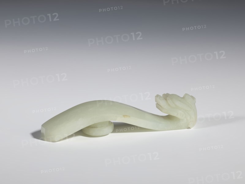 Belt clasp, stones; Chinese; dragons iconography; Qing style, Qing dynasty 1644–1911; milky-white jade with colloid clusters, full-plastic carved head; 18th–19th century, China; belt buckle; height 2.1 cm, width 10.1 cm, depth 2.2 cm., A smooth, pale green jade carving shaped like a curved object rests on a light surface, with a rounded support piece beneath its middle; one end tapers to a flattened tip while the other end features a carved floral or leafy motif with layered, rounded petals or leaves; soft shadows appear beneath and to the left of the object, and the background transitions from light gray near the surface to a darker gray toward the top.