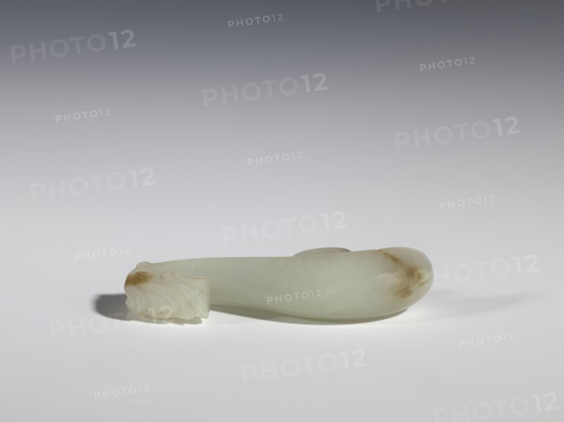 Belt clasp, Chinese, dragons iconography, Qing 1644–1911, milky-white jade with brown veins and clusters of colloids; full-sculpture carved head, engraved; 18th–19th century, China; belt buckle; height 1.9 cm, width 7.4 cm, depth 1.6 cm., A smooth, elongated object made of pale greenish-white stone with a few brownish discolorations rests horizontally on a flat, light-colored surface. The left end shows a carved, detailed section with layered, rounded forms and a small brown patch, while the right end tapers into a rounded, bulbous shape with a brownish area. Soft shadows fall beneath and to the left of the object, and the background transitions from light near the surface to darker gray toward the top.