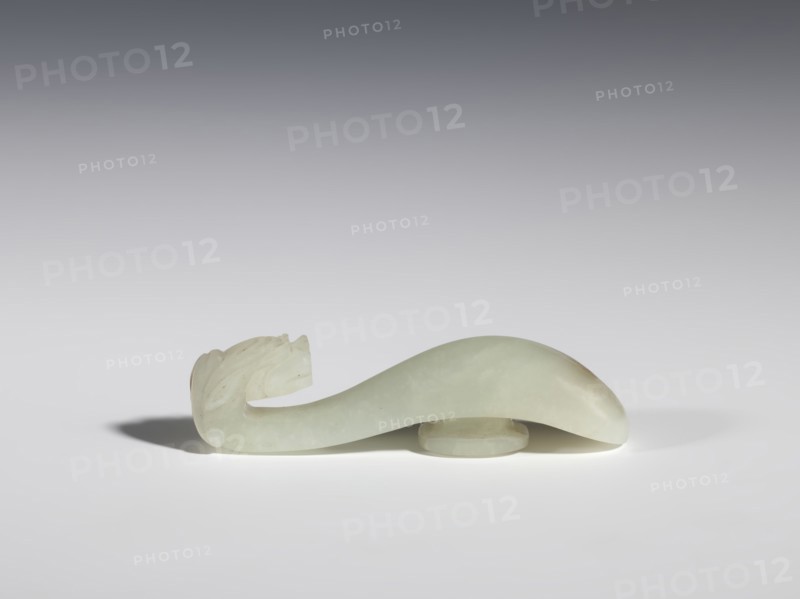 Belt clasp, Chinese, dragons iconography, Qing 1644–1911, milky-white jade with brown veins and clusters of colloids, full-sculpture carved head, engraved, 18th–19th century, China, belt buckle; height 1.9 cm, width 7.4 cm, depth 1.6 cm, A pale greenish-white carved object with a smooth, curved body and a carved end resembling a stylized head rests on a flat surface; the object casts a soft shadow and sits partly elevated on a small circular base. The background shows a smooth gradient from darker gray at the top to lighter gray near the surface, and the visible colors include pale green, white, gray, and subtle beige tones.