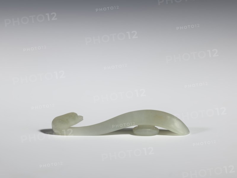 Belt clasp, Chinese, birds, Qing 1644–1911, milky-white jadeite with slight green tint, full-sculpture carved head, handle with an edge on the spine and a pointed tip, 17th–18th century, China, belt buckle; h. 1.3 cm, w. 7.3 cm, d. 0.96 cm., A smooth, curved object resembling a stylized snake or serpentine form rests on a flat surface, carved from a pale green, almost translucent stone with subtle variations in hue; one end rises slightly with a rounded head-like shape and a small open mouth, while the body arches gracefully and is supported near the center by a low, disc-like element, casting soft gray shadows on the white-gray background.