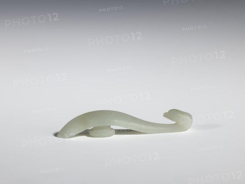 Belt clasp, Chinese, Chinese art; birds; Qing dynasty 1644–1911; 17th–18th century; milky-white jadeite with slightly green tint, clusters of colloids; full-sculpture carved head; handle with an edge on the spine and a pointed tip; belt buckle; height 1.3 cm, width 7.3 cm, depth 0.96 cm., A smooth pale green, translucent curved object resembling a stylized animal form rests on a pale gray surface; it has a rounded body that tapers into a slender neck and head with subtle carved features, supported near its underside by a small rounded base, and casts a soft shadow on the surface, with colors visible including pale green, light gray, and hints of white.