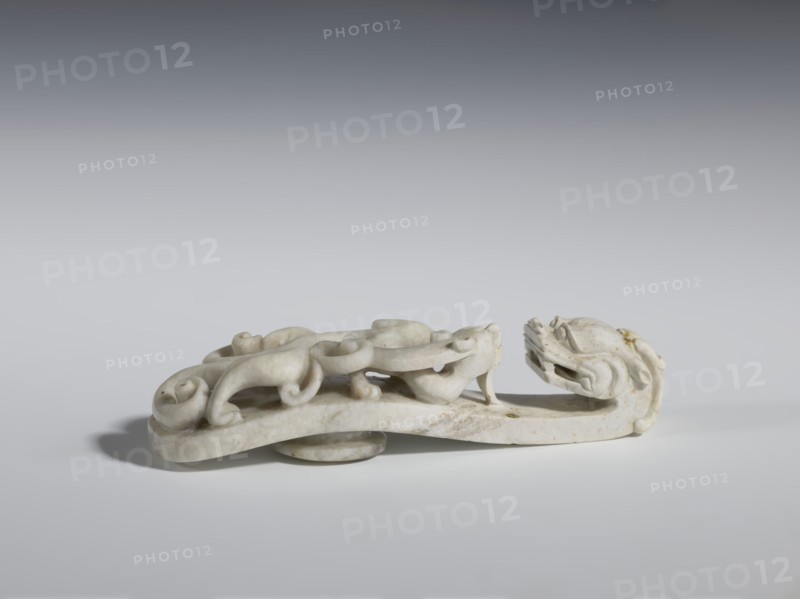 Belt clasp. Handle: stylized lizard-dragon qilong. Milky-white jadeite. Full-plastic carved head; full-plastic openwork handle. 18th–19th century, China. Belt buckle; height 2.8 cm, width 12.0 cm, depth 2.9 cm. Categories: Chinese art; Qing 1644–1911; men's clothing; costumes; ornaments; men's ornaments; lizards; dragons., Purchase, A low, elongated carved object with a stylized animal head at one end and a sinuous, scrolling body along its length, rendered in pale off-white stone with subtle areas of darker discoloration and small brownish spots; the piece sits on a flat surface casting a soft gray shadow and is photographed against a smooth gradient background ranging from light gray at the bottom to darker gray toward the top.