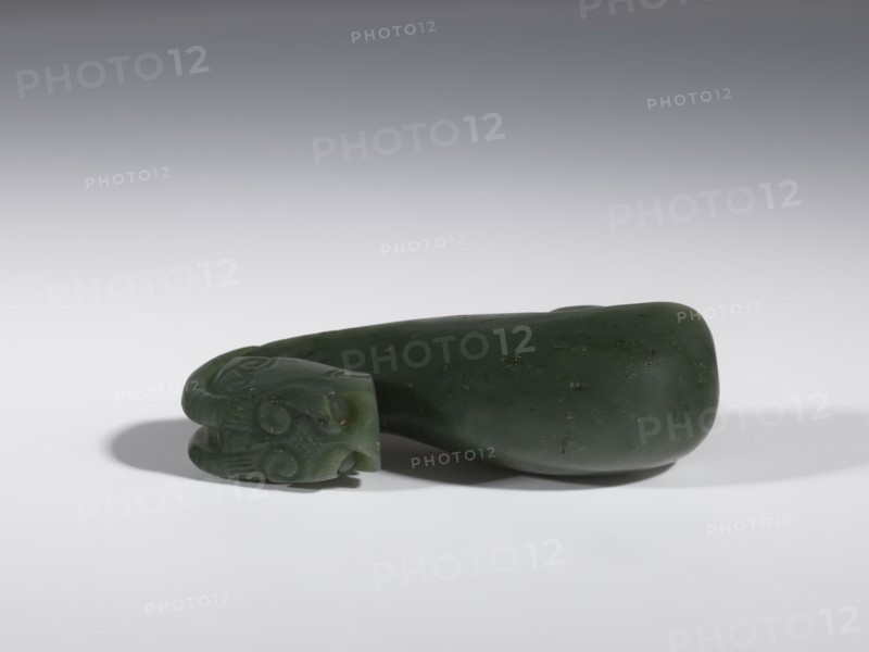 Belt clasp, Chinese, Chinese art, dragons iconography, Qing 1644–1911, clothing, belts, men's clothing, costumes, ornaments, men's ornaments, buckles, spinach jade, uniformly colored, full-plastic carved head, engraving, bas-relief, 18th–19th century, China, belt buckle; height 3.5 cm, width 9.5 cm, depth 3.1 cm, A smooth, dark green stone object rests on a light surface with a soft shadow beneath it; one end of the object features carved decorative swirls and rounded shapes, while the rest tapers into a polished, irregularly rounded form with small darker flecks; the background transitions from pale gray near the surface to a darker gray toward the top, and visible colors include shades of dark green, light gray, and darker gray.