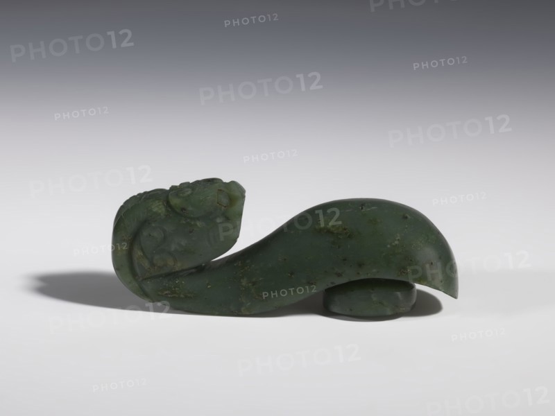 Belt clasp, purchased provenance, Chinese, dragons iconography, Qing 1644–1911, clothing, belts, men's clothing, costumes, ornaments, men's ornaments, buckles, spinach jade, uniformly colored, full-plastic carved head, engraving, bas-relief, 18th–19th century, China, belt buckle; height 3.5 cm, width 9.5 cm, depth 3.1 cm., A smooth carved object made of dark green stone with lighter green and brown mottling sits on a white surface against a gray gradient background; the carving features a curved, elongated body that rises into a rounded head with subtle carved facial and decorative details, and a small rounded base piece partially visible beneath the larger form, casting soft shadows on the surface.