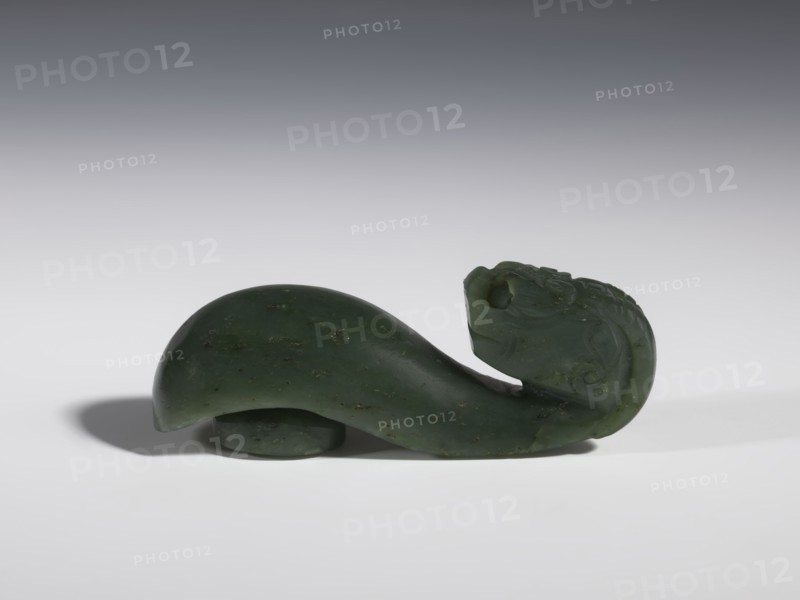 Belt clasp, Chinese, dragons iconography, Qing 1644–1911, men's clothing/ornaments, buckles, spinach jade, uniformly colored, full-plastic carved head, engraving, bas-relief, 18th–19th century, China, belt buckle; height 3.5 cm, width 9.5 cm, depth 3.1 cm., A carved object in dark green with lighter green and small brownish flecks rests on a light surface against a gradient gray background; the object has a smooth, curved body that rises and tapers toward one end where a carved head with rounded features and shallow carved patterns is visible, and a small rounded support piece sits beneath the body, casting a soft shadow on the surface.
