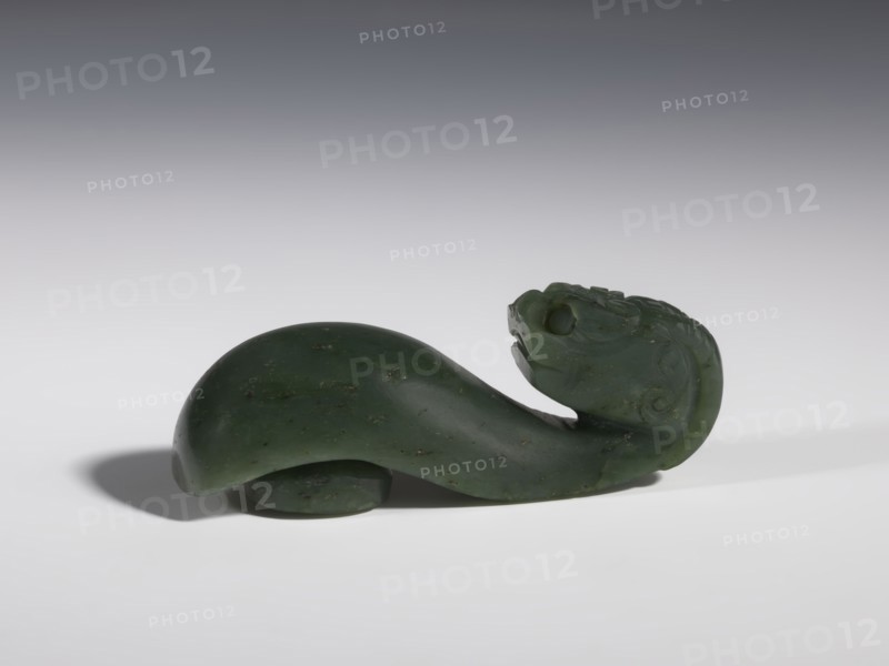 Belt clasp, Chinese, dragons iconography, Qing 1644–1911, clothing, belts, men's clothing, costumes, ornaments, men's ornaments, buckles, spinach jade, uniformly colored, full-plastic carved head, engraving, bas-relief, 18th–19th century, China, belt buckle, height 3.5 cm, width 9.5 cm, depth 3.1 cm, A carved dark green stone object shaped like a stylized animal form with a curved body and an upturned head, showing carved facial features and surface detailing; the stone has small darker flecks and a slightly glossy finish, and the piece rests on a smooth light-gray surface with a soft shadow beneath and a gradient gray background.