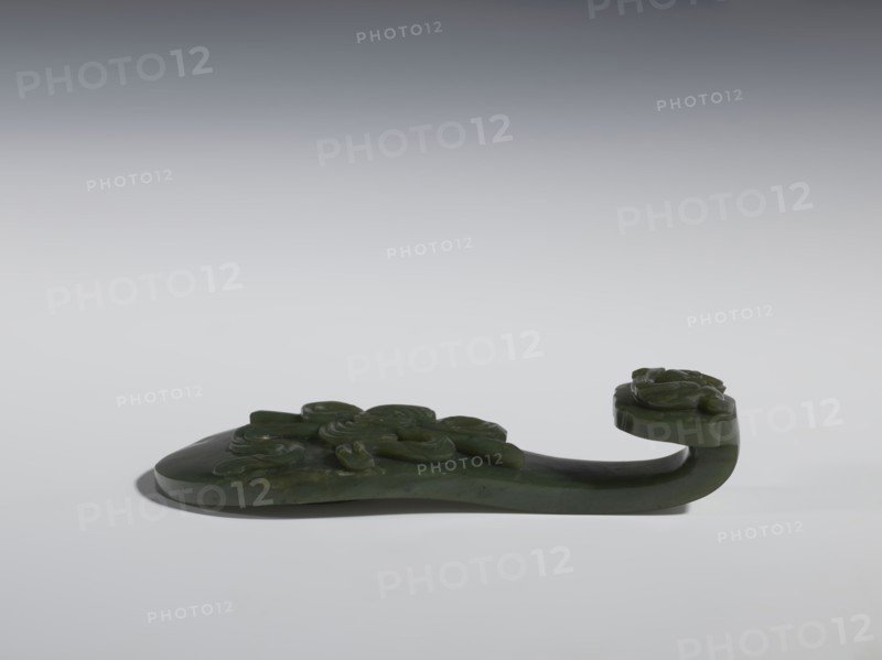 Belt clasp. Scepter head with a bas-relief stylized lizard; shaft with a Taoist emblem of one of the Eight Immortals. Spinach jade. Sculpture, engraving, 18th–19th century China. Chinese art. Chinese culture. Taoism. Qing 1644–1911. Subject: lizards; ruyi auspicious scepter; Immortals. Belt buckle. Height 2.7 cm; width 12.0 cm; depth 4.5 cm., Purchase provenance., A carved dark green object shaped with a flat rounded base and a curved raised handle, the surface decorated with carved floral motifs and rounded petal forms, shown against a smooth gradient gray background with soft shadows beneath; visible colors include dark green, lighter green highlights, gray, and white.