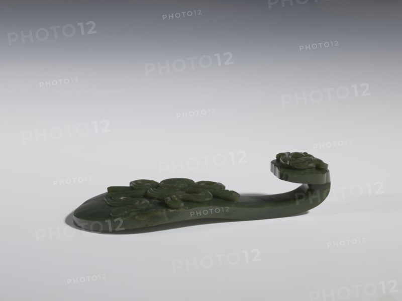 Belt clasp, Chinese, Qing 1644–1911, 18th–19th century. Spinach jade; sculpture and engraving. Scepter head with a bas-relief stylized lizard; shaft with a Taoist emblem of one of the Eight Immortals and ruyi auspicious scepter iconography; Immortals and Taoism themes; lizards zoological motif. Belt buckle. Height 2.7 cm; width 12.0 cm; depth 4.5 cm., A carved object made of dark green material rests on a light surface with a gradient gray background, showing smooth and glossy finishes and a few areas of subtle translucence; the piece has an elongated base with raised carved floral motifs and a curved extension that rises and ends in a rounded platform also decorated with carved flower shapes, casting a soft shadow beneath.