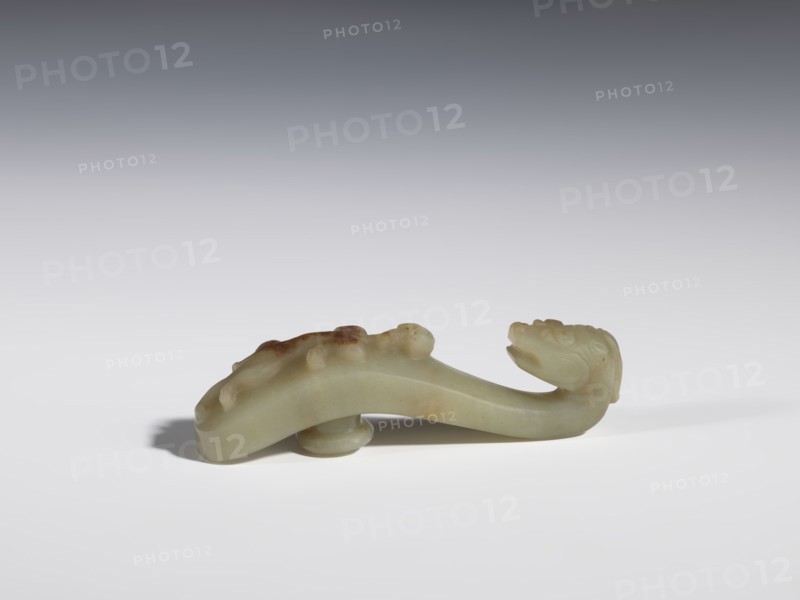 Belt clasp. Handle flattened on the sides, decorated with a lizard-dragon motif. Jadeite with traces of chalcedony. Head carved in full plastic, convex relief. Dragons; lizards. Chinese, Qing 1644–1911. 17th–19th century. Belt buckle. Height 2.3 cm; width 8.4 cm; depth 1.8 cm., A smooth sculpted object resembling a curved creature with a pronounced head at one end and a tapering body, carved from a pale greenish material with areas of darker brownish discoloration, resting on a flat white surface with a soft shadow beneath; the background fades from light gray near the surface to a darker gray toward the top, and the carving shows rounded raised details along its back and a small rounded support under its midsection.