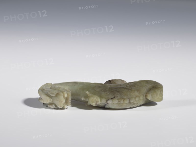 Belt clasp;, Chinese, Qing dynasty 1644–1911, 19th century. Material not specified. Head in the shape of a two-horned dragon's head with an open mouth; shaft carved as a dragon playing with a flaming pearl. Head and shaft carved in full plastic. Function: belt buckle for men's clothing/ornament. Iconography: dragons, dragon with pearl. Dimensions: height 2.65 cm; width 9.7 cm; depth 1.9 cm., A carved object rests on a flat surface, appearing to be made of a smooth, translucent material in shades of pale green, olive, and hints of brown, with variations and darker veins. The object has a curved, elongated form with rounded ends and a low, raised central area; one end features a carved motif that resembles a stylized animal head with curved, layered shapes, while the body shows a row of rounded protrusions and subtle carved lines. The surface catches soft light, creating gentle highlights and shadows, and there is a diffuse gray-to-white background behind the object.