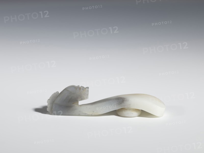 Belt clasp, Chinese, dragons iconography, Qing 1644–1911, flat profiled handle. White jadeite with gray shading, fully carved head. 17th–19th century, China. Belt buckle; H 2.1 cm, W 8.9 cm, D 2.0 cm., A smooth carved object rests on a flat surface, showing an elongated curving form with a rounded end and a carved head-like shape at the other end with ridged details; the object appears to be made of a translucent stone with areas of white, pale gray, and subtle beige, and it casts a soft shadow on the surface beneath it.