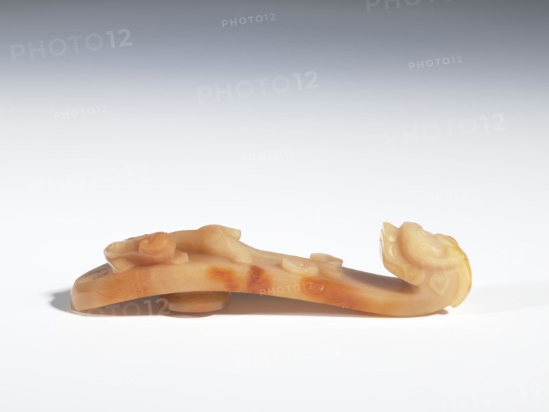 Belt clasp, Chinese, Chinese art; tigers, dragons; Qing 1644–1911. On the handle are engraved patterns in the shape of bats, a ruyi scepter head, and a leafy flower branch. Beige jadeite with darker spots. Carving, bas-relief, engraving; 18th–19th century, China. Belt buckle; height 2.4 cm, width 11.0 cm, depth 2.4 cm., A carved object made of a translucent warm beige to honey-colored material rests on a white surface, showing areas of darker amber-orange mottling; the piece has smooth, rounded forms with a curled end shaped like an animal head and raised decorative bumps along its top, casting a soft shadow beneath.
