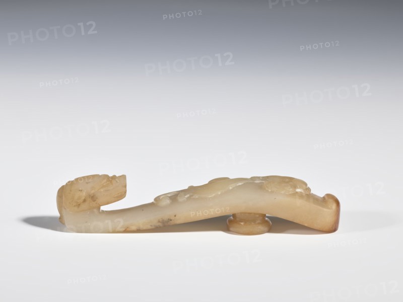 Belt clasp, Chinese, dragons iconography, Qing 1644–1911, bas-relief handle with a projection at the bottom — lizard-dragon chilong; head carved in full plastic. Jadeite with chalcedony inclusions. 17th–19th century, China. Belt buckle. Height 2.1 cm, width 10.6 cm, depth 1.8 cm., A carved object made of a pale, creamy stone with areas of light brown and amber tones lies horizontally on a flat white surface against a light gray gradient background; the object has a rounded raised base near one end and a curved elongated body with subtle carved bumps and shallow grooves, one end rising into a stylized head-like shape with carved facial features, and the opposite end curling slightly upward with a darker amber edge.