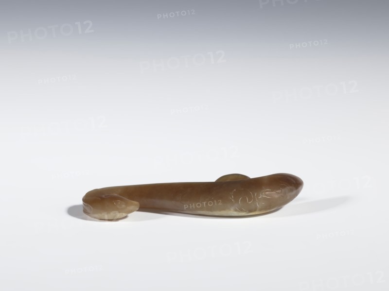 Belt clasp, Chinese, Qing 1644–1911, 18th–19th century. Brown jade with lighter spots, full-plastic carved head, engraved. Subject: birds, phoenixes, dragons iconography. Function: belt buckle. Dimensions: height 1.6 cm, width 7.4 cm, depth 1.2 cm., A smooth, elongated object rests on a white surface, with a rounded end resembling a small head and a thicker curved opposite end; the object is primarily brown with lighter tan areas and subtle pale greenish-yellow tones, and faint carved or etched swirling patterns are visible along one side, casting soft gray shadows on the surface.