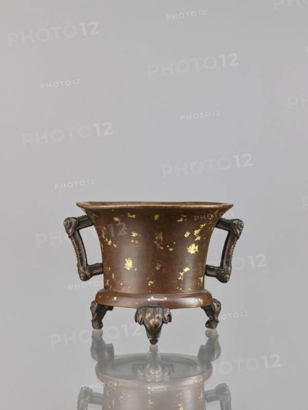 Censer, Chinese, Qing dynasty 1644–1911, 18th century. Bronze ritual/ceremonial vessel censer with elephant motifs; backdated signatures. Dimensions: height 9.7 cm, width 11.3 cm., A small metal vessel with a flared rim and a rounded body sits on ornate legs, showing a brown surface with scattered gold patches; it has a pair of decorative handles with curled ends and sculpted detailing, and the three legs terminate in stylized animal-like feet, all reflected faintly on a smooth gray surface beneath; the background is a uniform light gray.