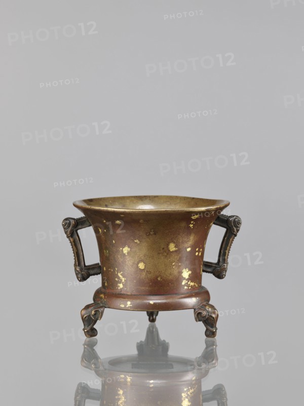 Censer, Chinese, Qing 1644–1911, 18th century, China; ceremonial/ritual bronze vessel with backdated signatures, decorated with elephants; height 9.7 cm, width 11.3 cm., A small metal vessel with a flared rim and a rounded body sits on a reflective surface; its surface shows patches of worn gilding giving gold flecks over a brown and bronze base color. The vessel has a pair of ornate handles on opposite sides, each handle dark bronze with decorative scrollwork, and it stands on three short, sculpted legs that curve outward and have darker bronze tones. The interior of the vessel appears smoother and more golden than the exterior, and the reflection beneath echoes the shape and colors of the metal object. The background is a smooth, even light gray.