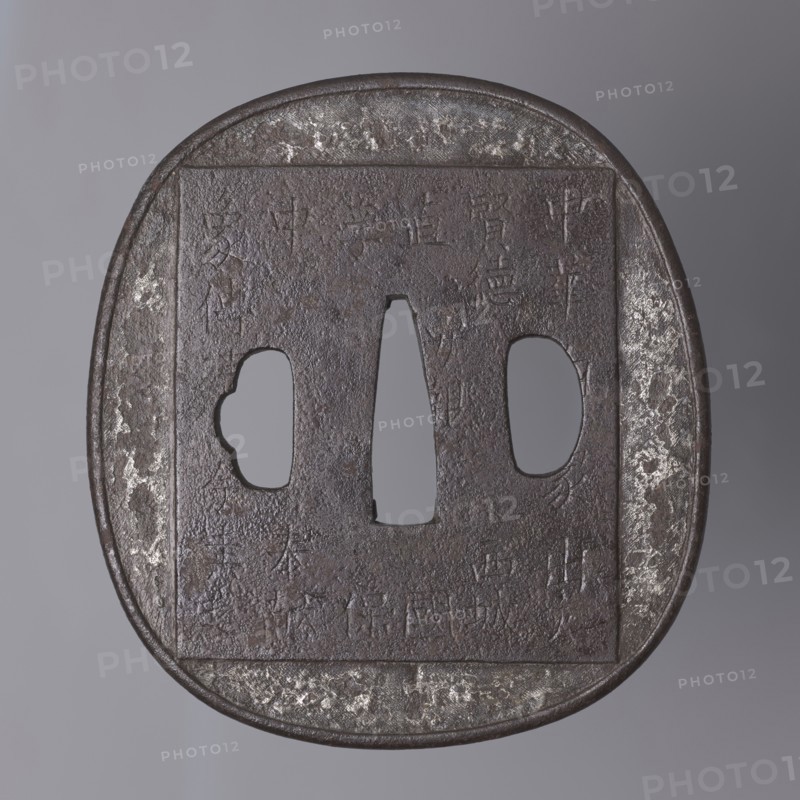 Crossguard tsuba, Tsuchiya Yasuchika 1670–1744, Nara school. Tsuba in the shape of a rectangle with rounded sides nadegaku gata, with a slightly distinct and rounded edge uchi kaeshi. On the obverse, low relief image of an elephant covering almost the entire surface; on the animal's back a saddle cloth decorated with a rosette and tendril border with a meander, and a Buddhist flaming jewel Cintamani. On the reverse a large rectangle with an inscription in kanji — a fragment of the Lotus Sutra; the backside bears a lengthy inscription relating to the arrival of the elephant in Japan. 18th/19th century Edo period, 1603–1868, Japan. Tsuba; height 8.7 cm, width 7.9 cm, depth 0.4 cm., A close-up view of a square-edged, rounded metal object with a central vertical opening flanked by two irregularly shaped side openings, showing a textured, pitted surface. The object displays a dark brown to almost black patina with areas of lighter gray and silvery patches where the surface appears worn. The...